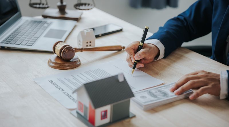 Image of someone sat at a desk writing on paper with a court gavel, weighing scale and model house also on the desk to support construction legislation article