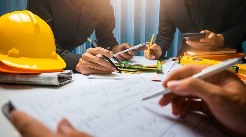 Image of a hard hat on a desk surrounded by documents and people writing notes to support construction management article